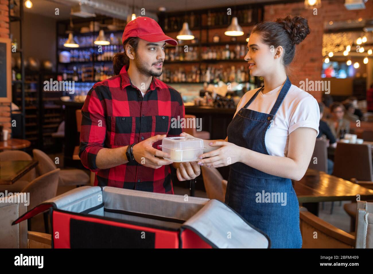 Young waitress in workwear passing plastic container with fresh food to courier while helping him to put orders of clients into big bag Stock Photo