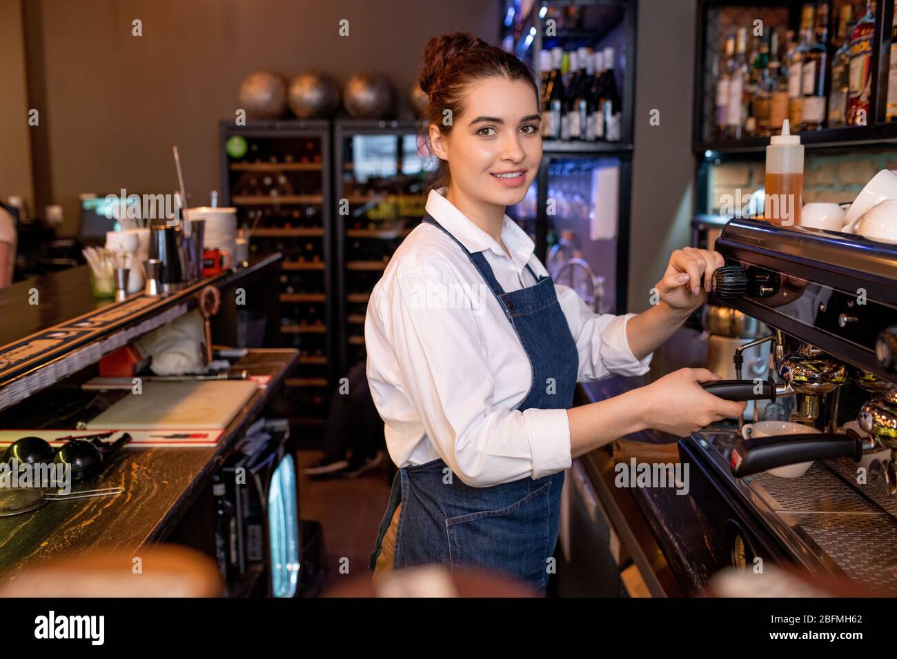 Pretty young friendly waitress in workwear preparing cappuccino while standing by coffee machine in cafeteria or restaurant Stock Photo