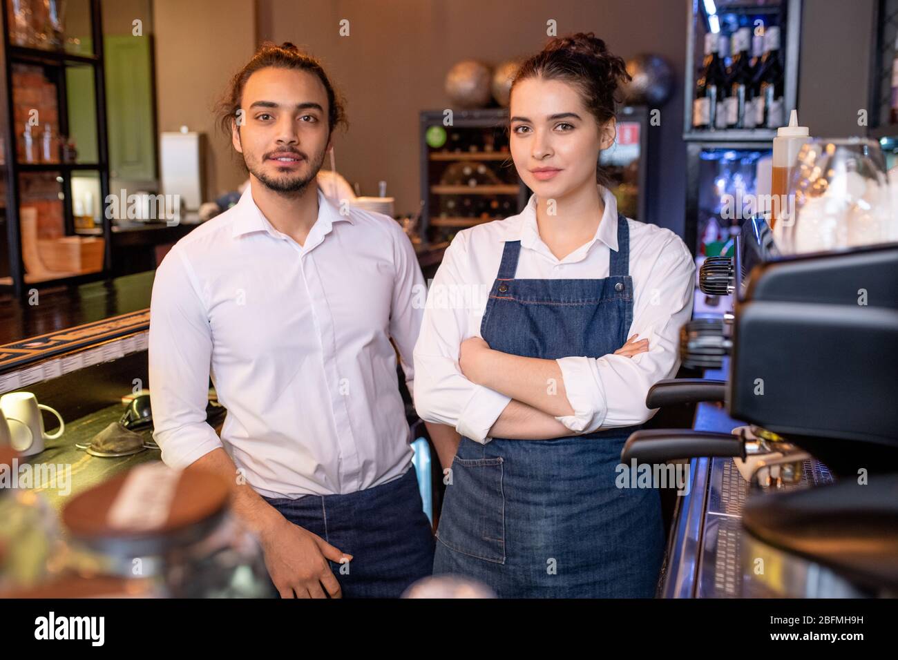 Two young colleagues in white shirts and denim aprons standing by workplace next to coffee machine in cafe and looking at you Stock Photo