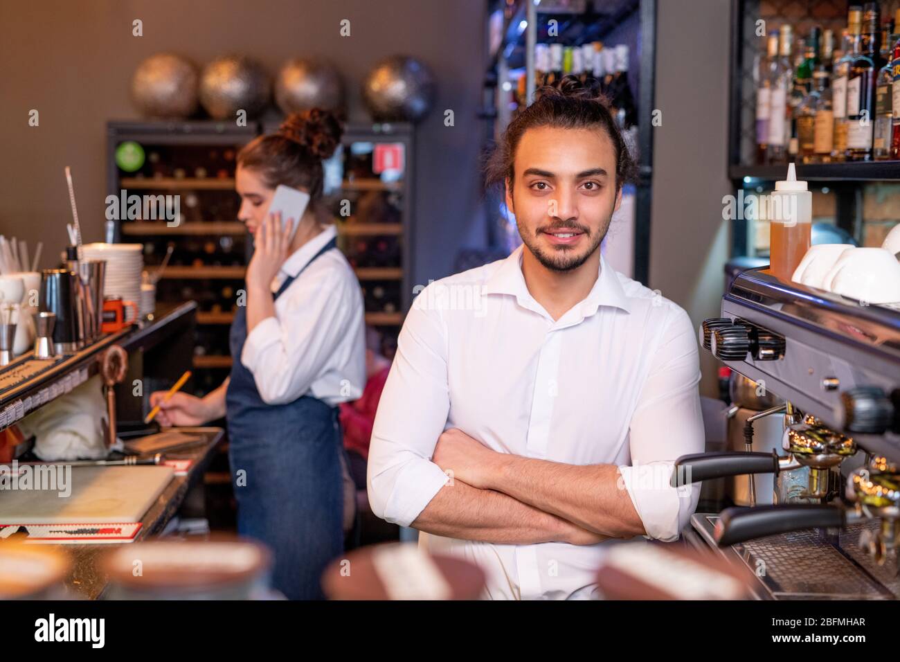 Young handsome waiter in white shirt crossing arms by chest while standing by coffee machine on background of colleague phoning Stock Photo