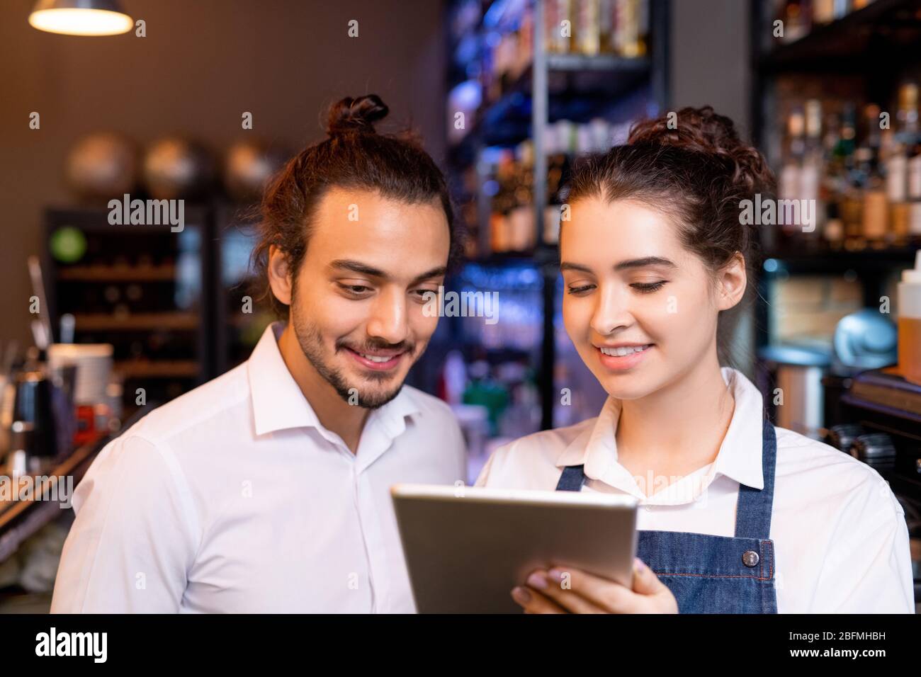 Happy young waitress with tablet showing her colleague new points in menu while discussing online orders of clients by workplace Stock Photo