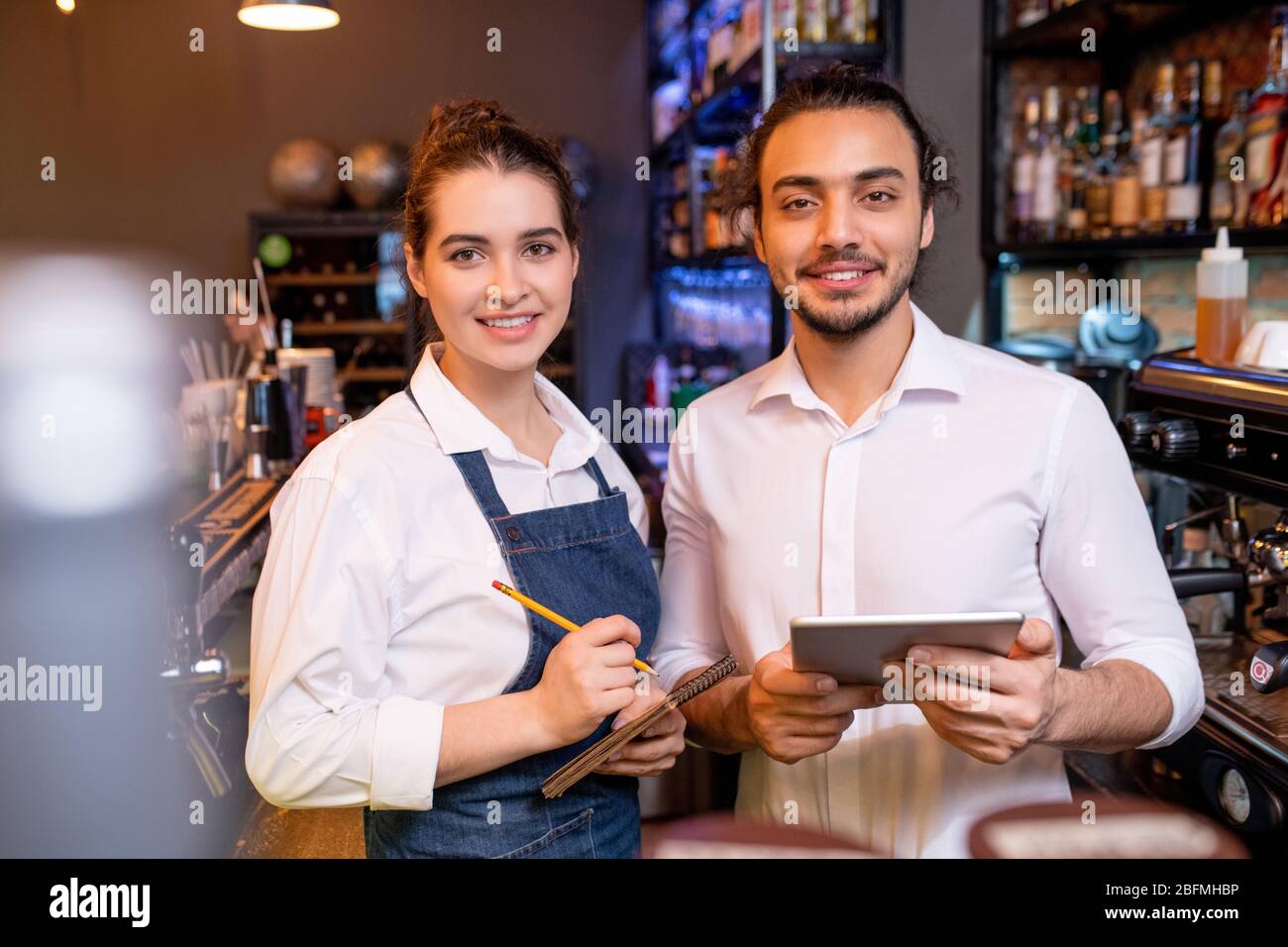 Two young cheerful workers of classy restaurant or cafeteria standing by workplace in front of camera and meeting new guests Stock Photo