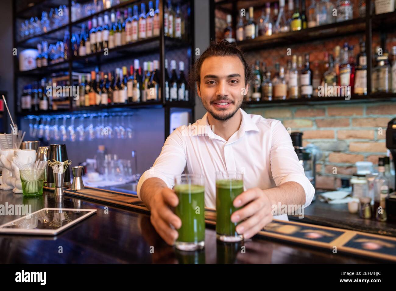 Happy young waiter or barman in white shirt passing you two glasses of fresh vegetable smoothie with bottles of alcohol on background Stock Photo