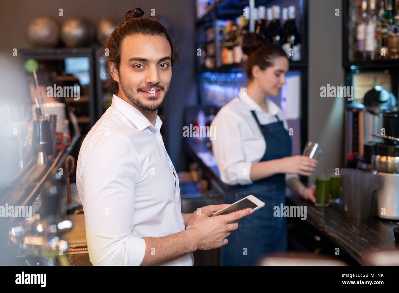 Young friendly waiter in white shirt scrolling through online menu while checking prices for drinks and food on background of colleague Stock Photo