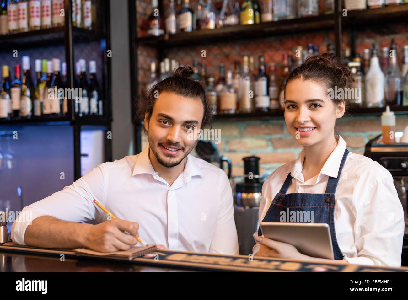 Happy young waiter making notes in notepad while his pretty colleague with touchpad standing near by and both looking at you Stock Photo