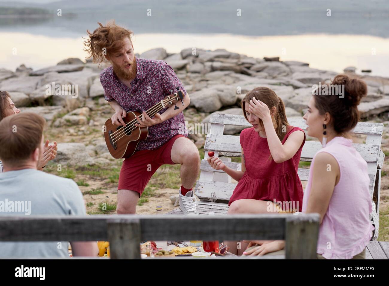 Young emotional bearded man playing the guitar while hanging out with friends outdoors near lake on warm summer evening Stock Photo