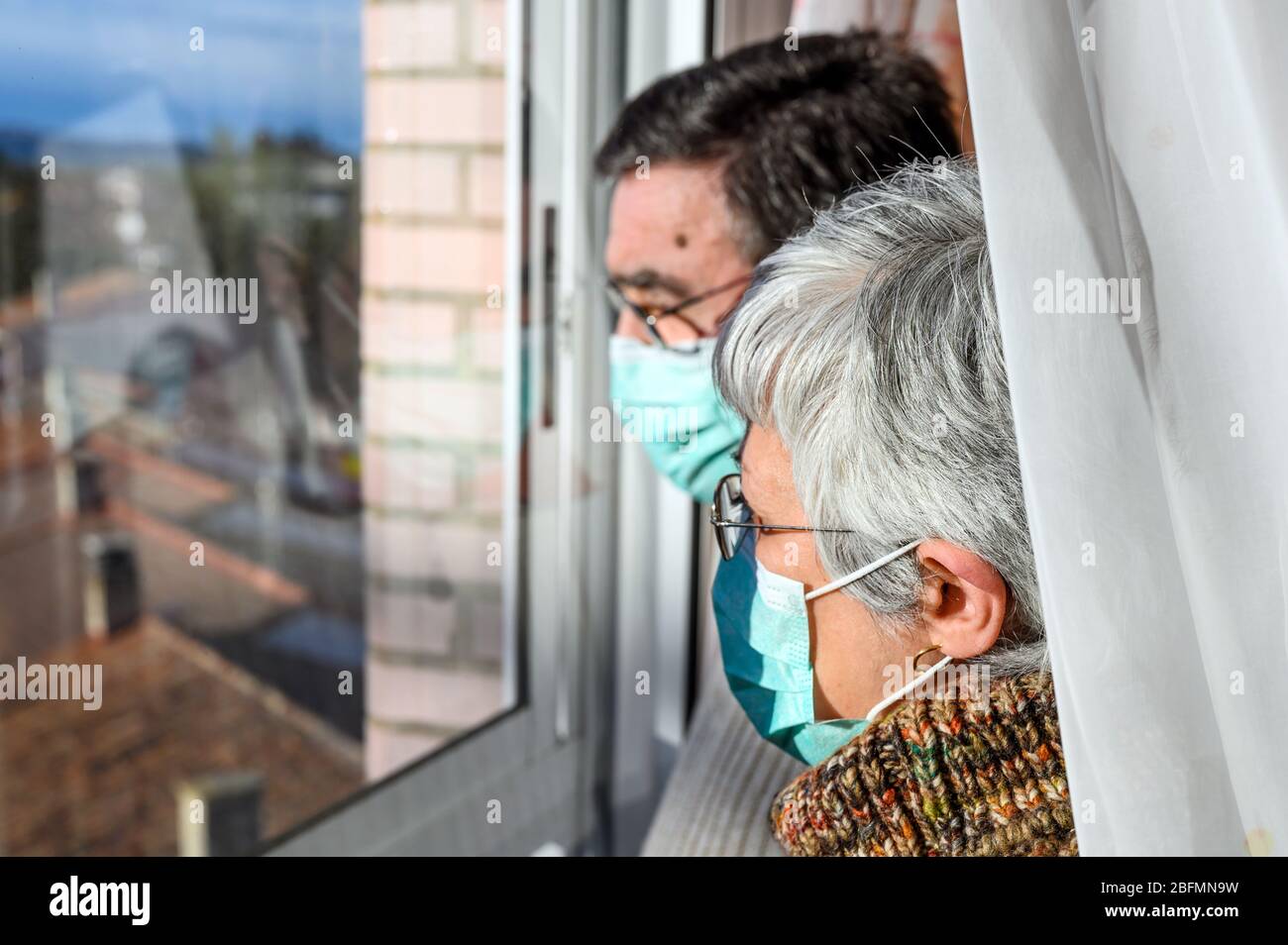 senior couple, with protective face masks, at home looking through the window. Concept of coronavirus quarantine stay home and social distancing. Lockdown people. elderly and retired lifestyle . Stock Photo