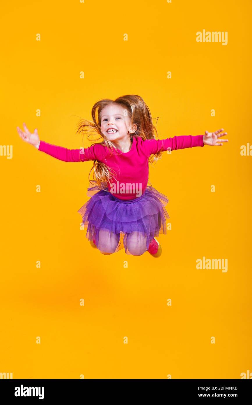Full length adorable little girl in colorful clothes smiling and jumping on yellow background Stock Photo