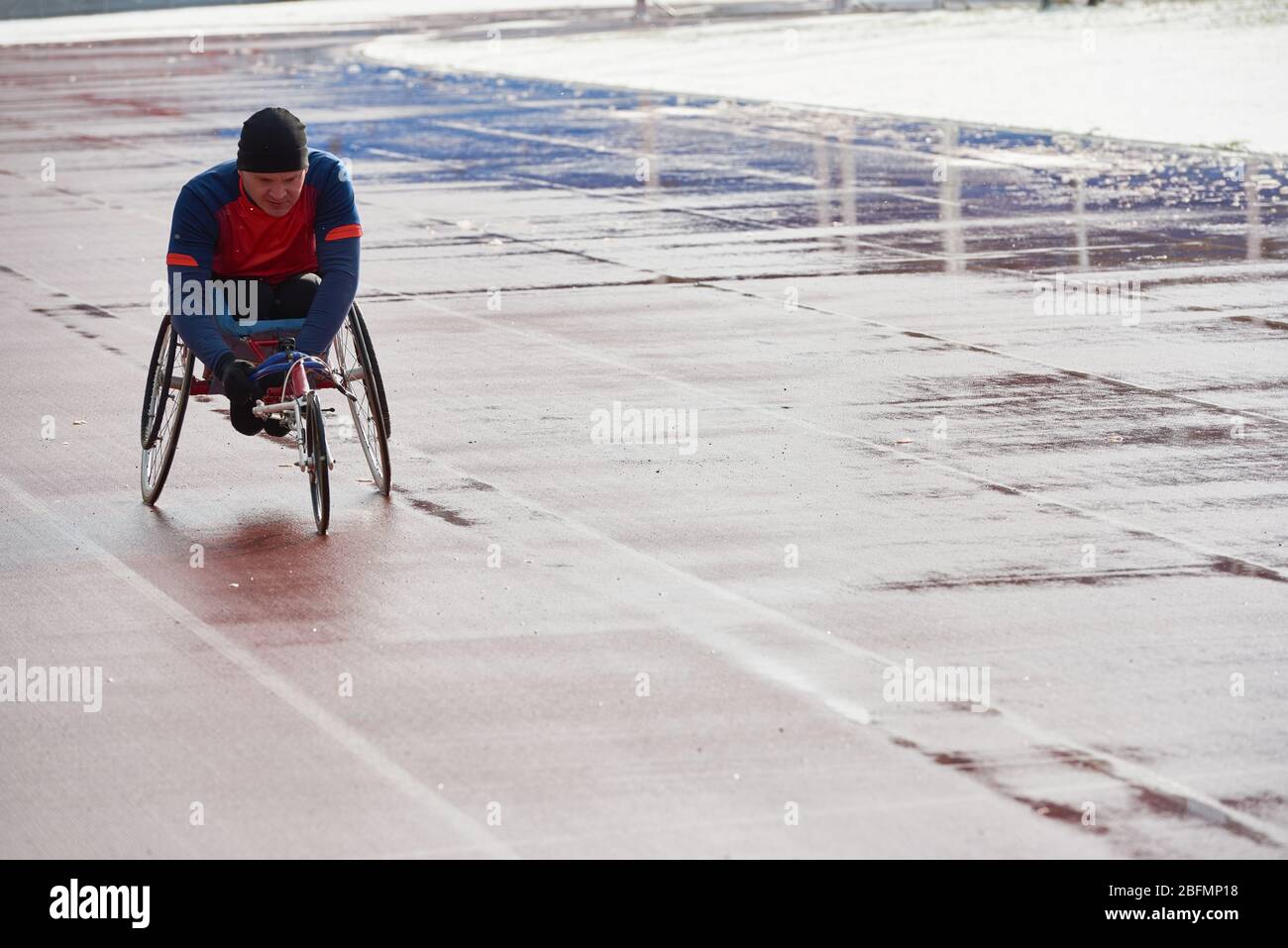 Wheelchair racing. Strong-willed handicapped male athlete in sport wheelchair training at outdoor track and field stadium on cold rainy day Stock Photo