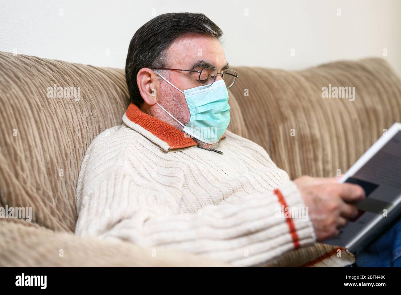 Covid- 19. Elderly man with protective face mask reading a book, while quarantine at home . Stock Photo