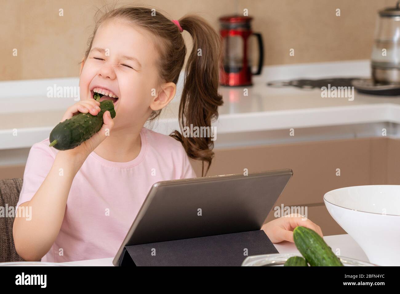 Little girl eating cucumber and using tablet at table in kitchen Stock