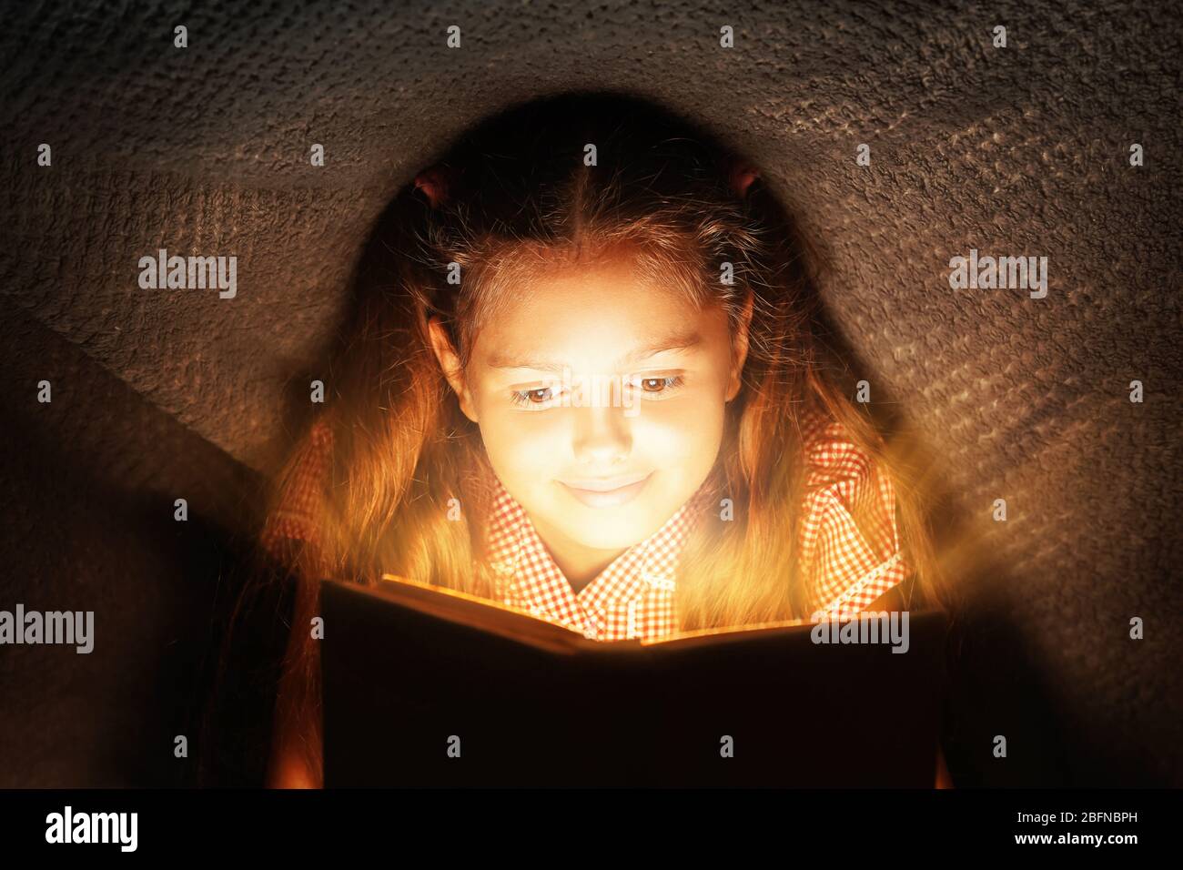 Cute girl reading book under blanket at night. Magic light coming out