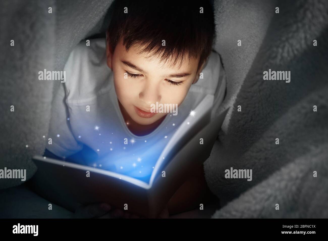 Cute boy reading book under blanket at night. Magic light coming out of