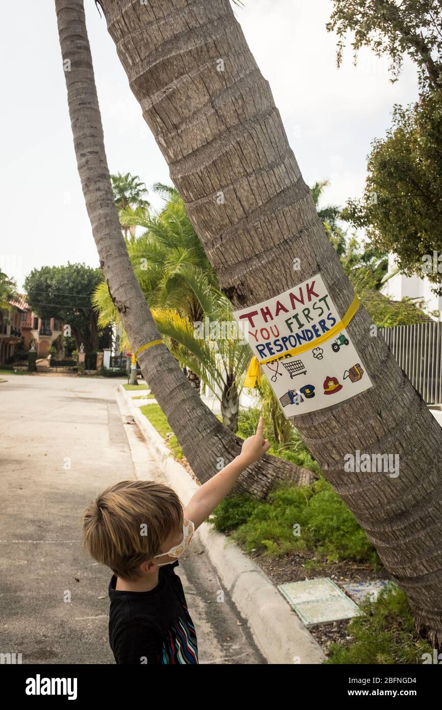 A young boy wearing a mask while out for a walk, looks at a home made sign stuck to a palm tree thanking first responders during the COVID-19 lockdown in Miami, Florida, USA. Stock Photo