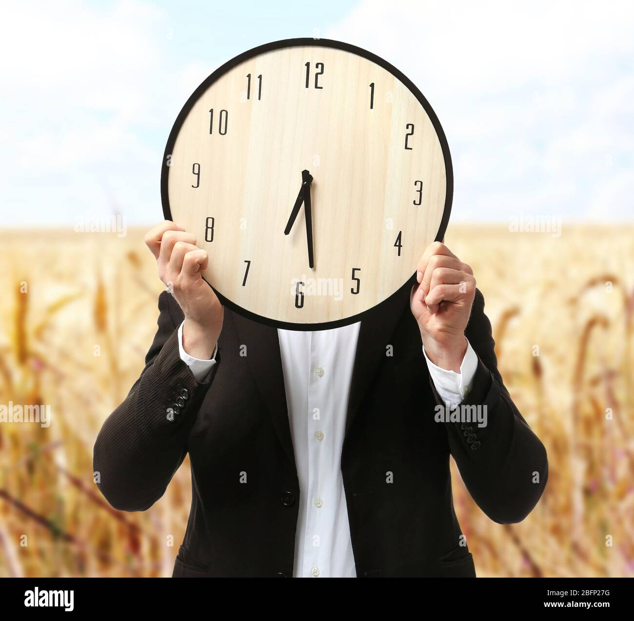 Man in black suit covering his face with big clock on blurred field