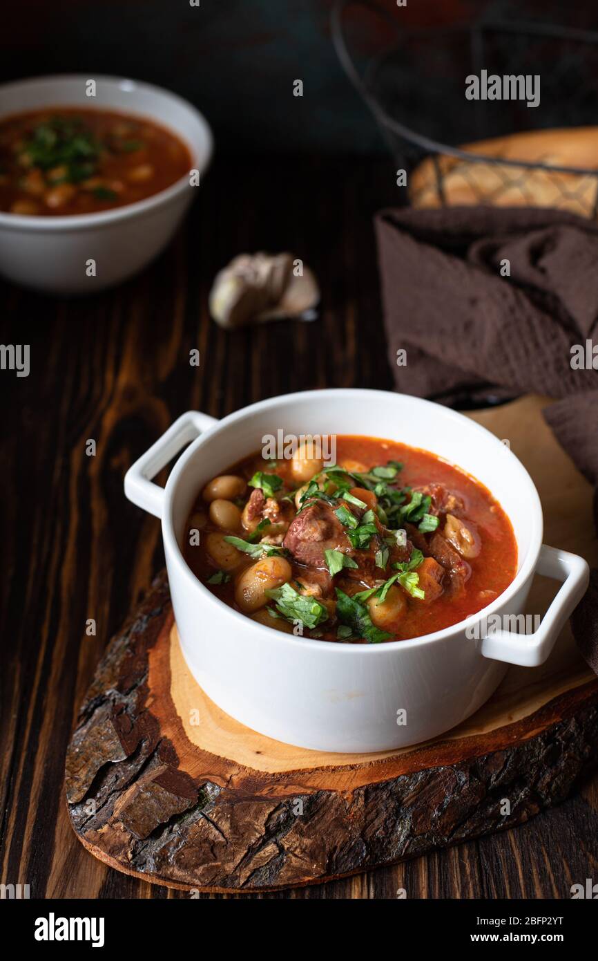 Bean soup with meat and vegetables served on a rustic board and wooden table with bread and garlic. Traditional Balkan soup Pasulj (Grah). Close-up Stock Photo