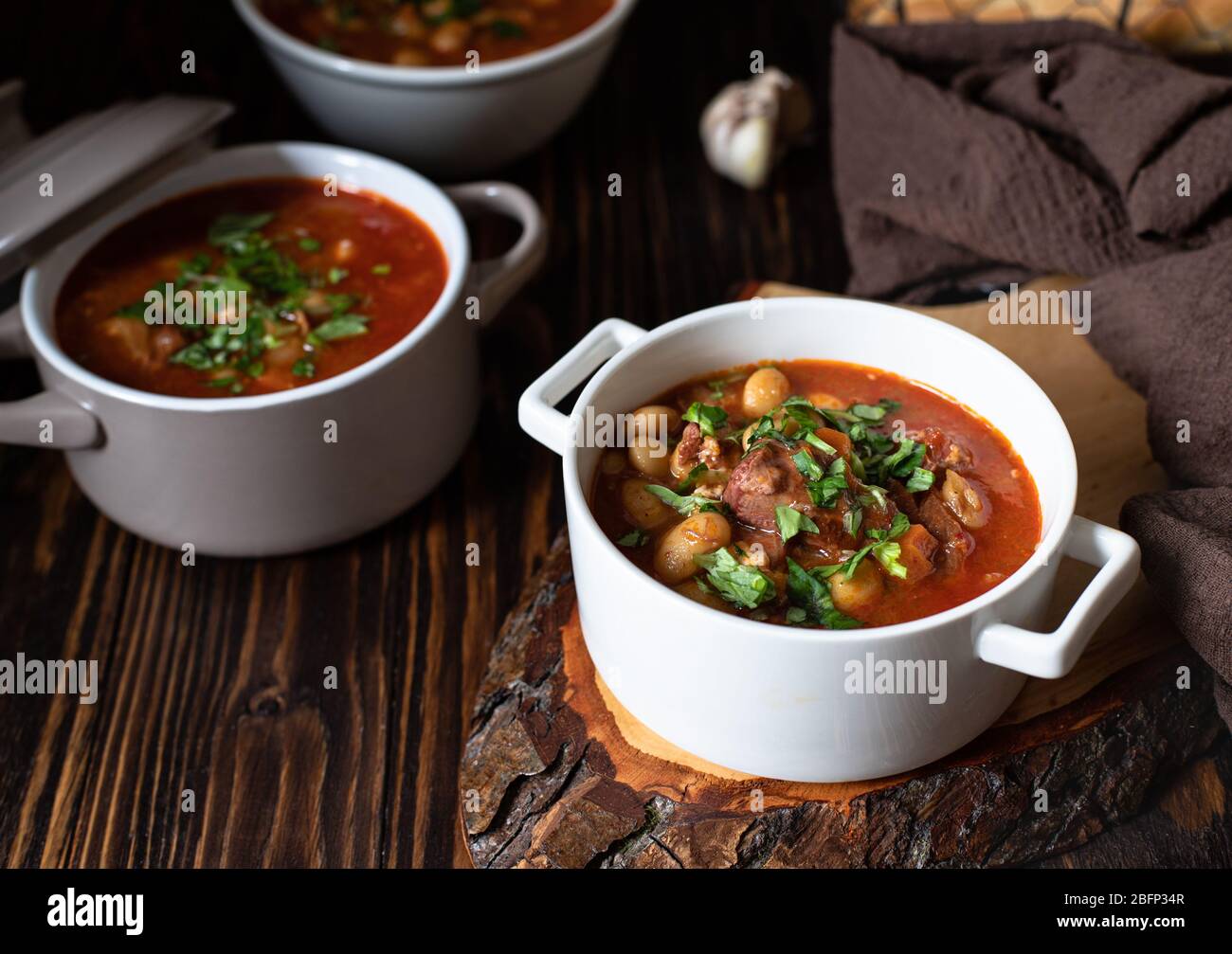 Bean soup with meat and vegetables served on a rustic board and wooden table with bread and garlic. Traditional Balkan soup Pasulj (Grah). Close-up Stock Photo
