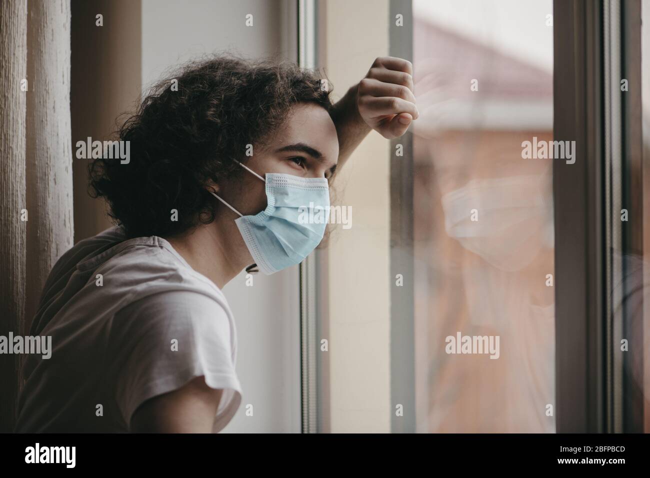 Teenager in protective medical mask looking out his home window. Quarantine concept. Stock Photo