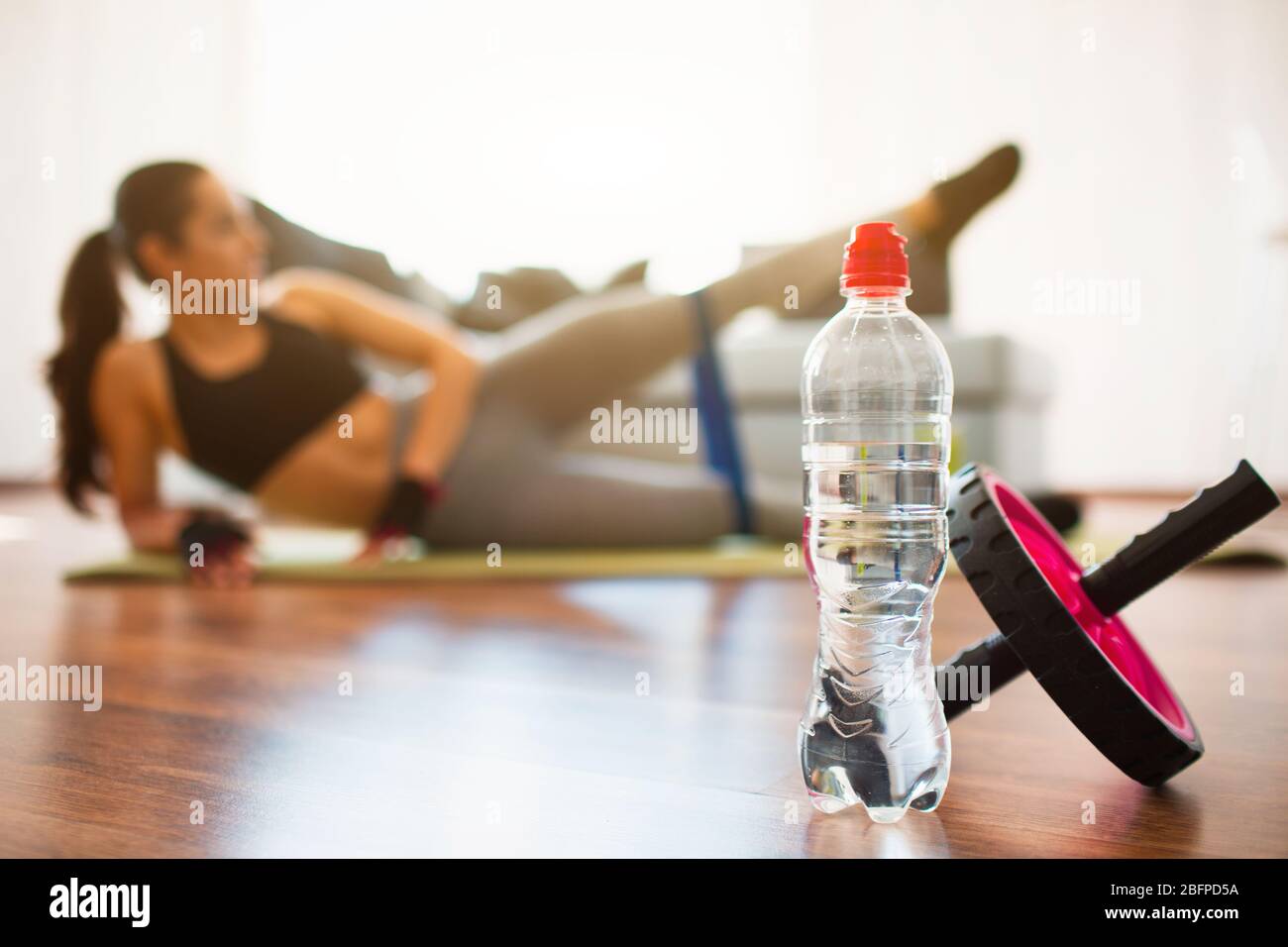 Young woman doing sport workout in room during quarantine. Water bottle and abdominal exercise roller in front. Girl stretching and training with Stock Photo