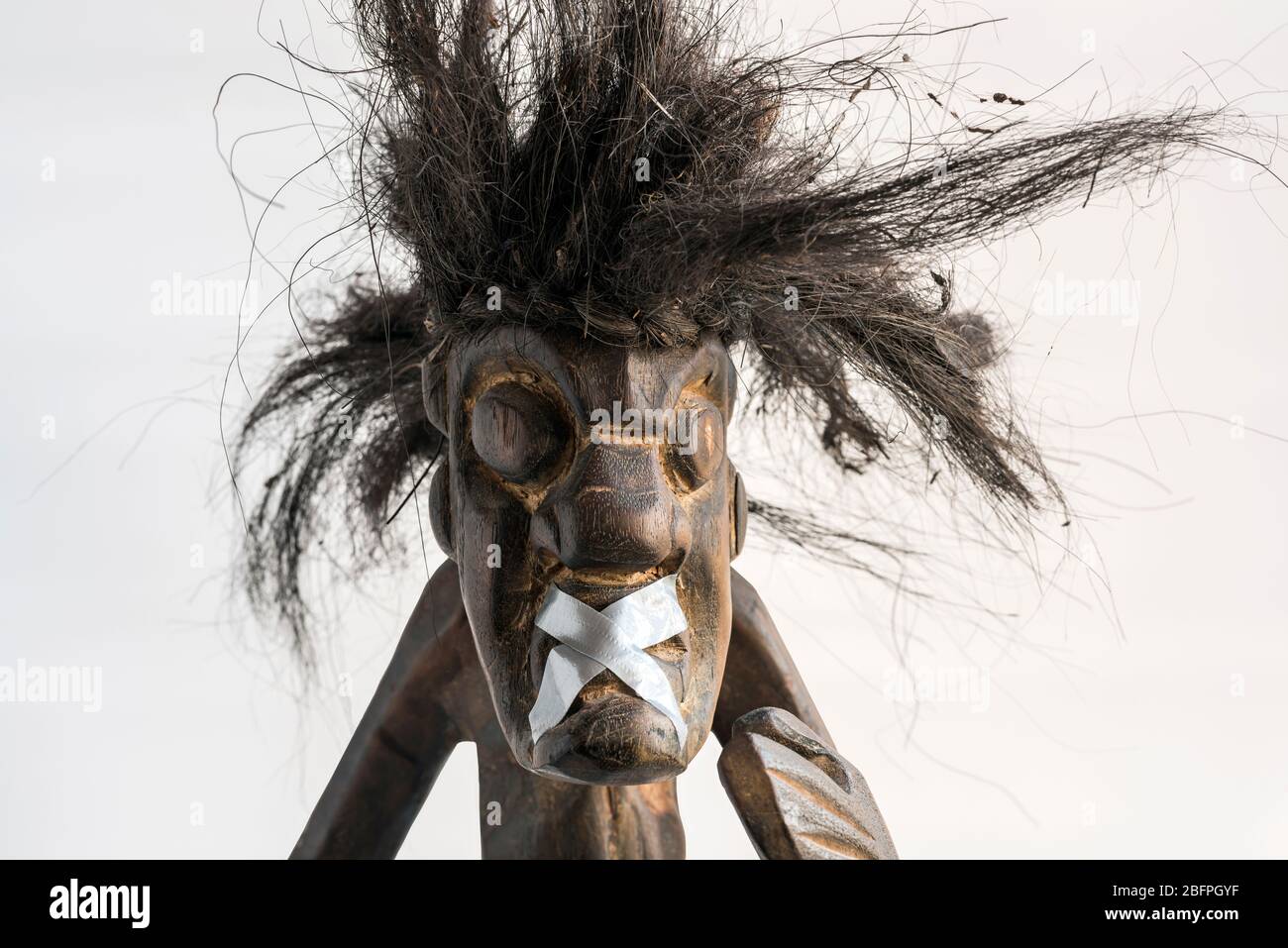 Wooden carved figure of primitive man wearing straw skirt & long black tousled hair, taped up mouth. Concept; Social immobility, silenced, ignored. Stock Photo