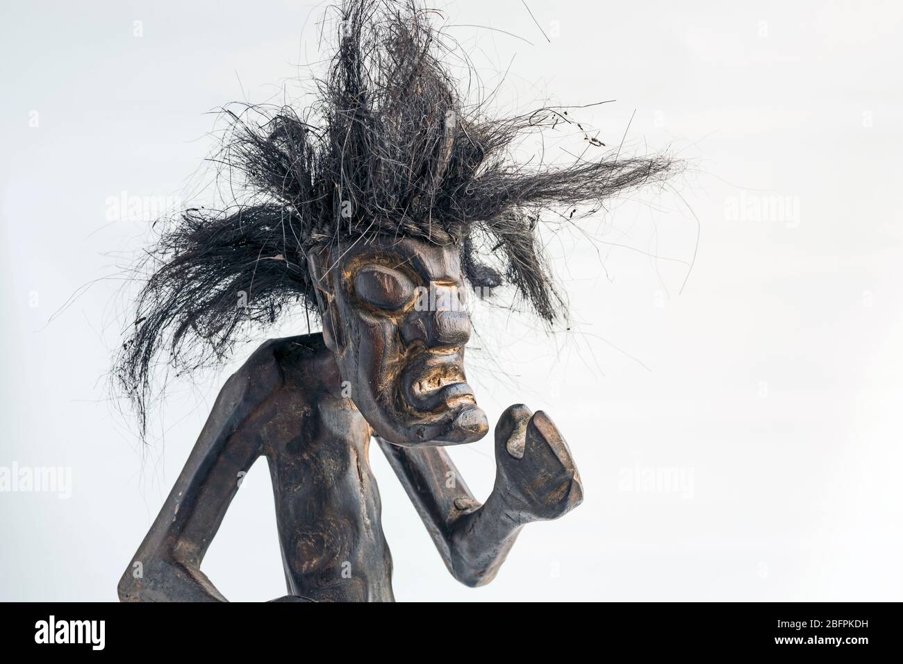 Wooden carved figure of primitive man wearing straw skirt & long black tousled hair. Concept; Bad hair day, difference, native, wild, unkempt, scary. Stock Photo