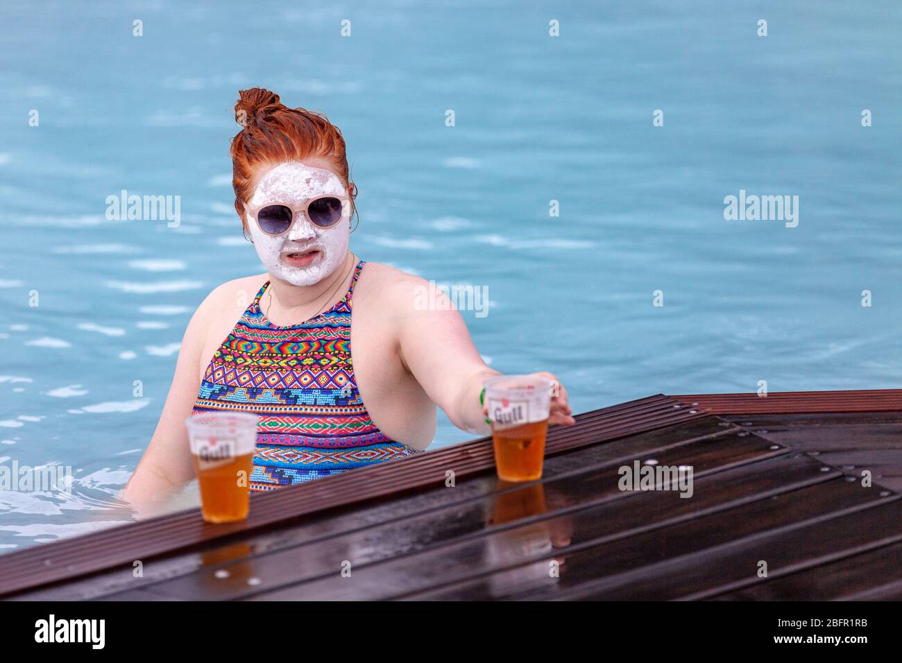 A woman wearing a mud face mask drinks beer at the Blue Lagoon Geothermal Spa Pool near Reykjavik in Iceland on a cold day in winter Stock Photo