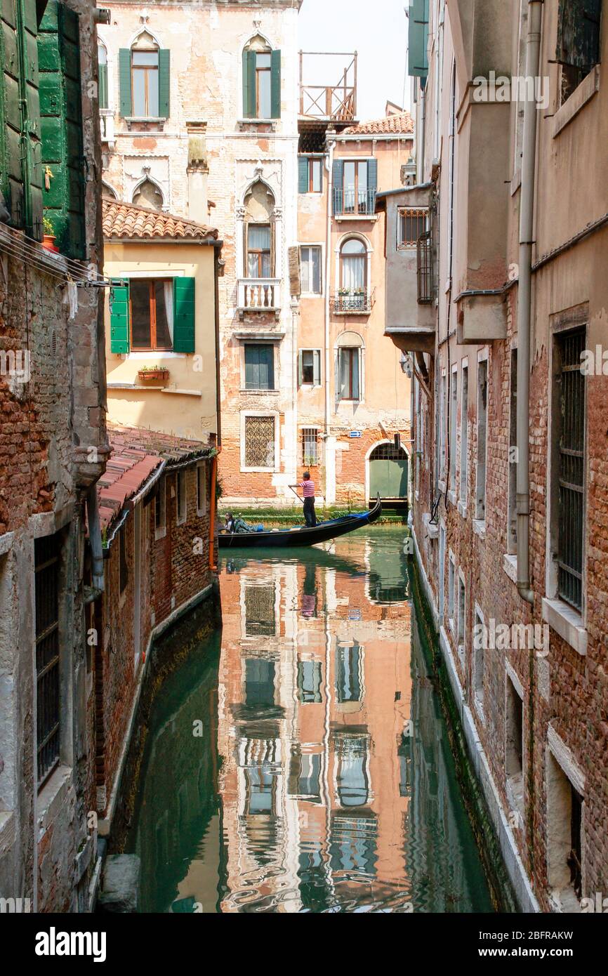 A gondola passing by a small side canal with reflections of buildings shown clearly. Venice, Italy Stock Photo