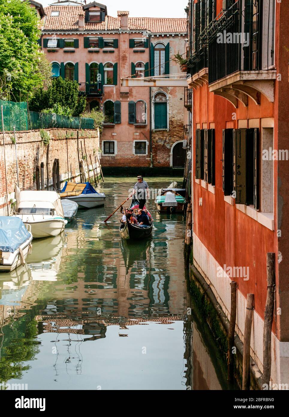 A gondola with people comes slowly in a small side canal surrounded by buildings. NMR Stock Photo