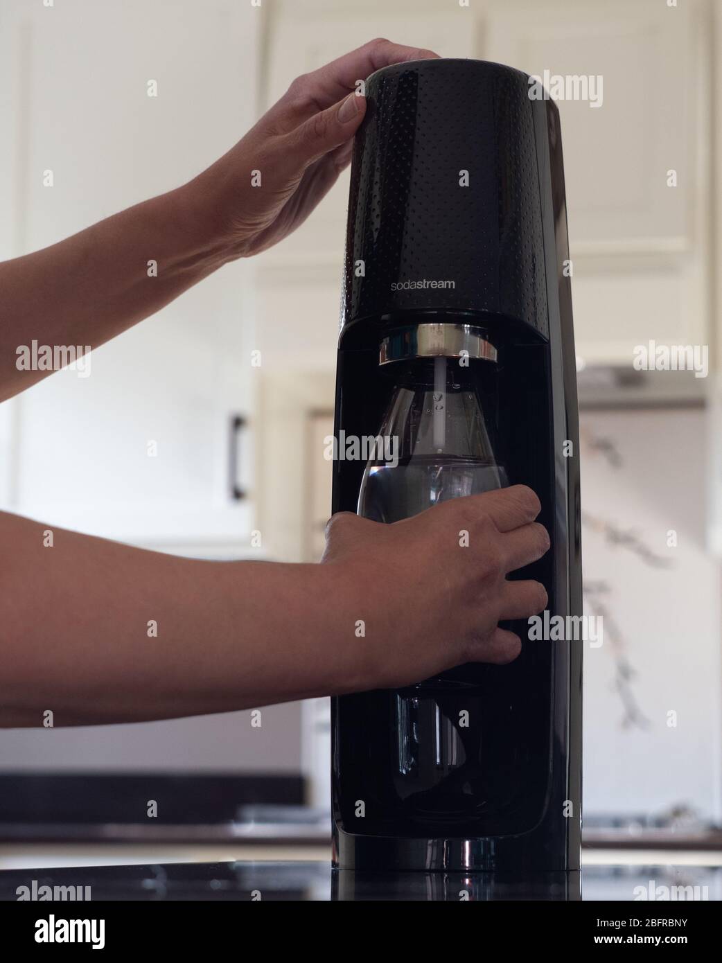 Latina woman using the Sodastream to carbonate water in a reusable plastic bottle in a modern kitchen with off white cabinets in the background. Stock Photo