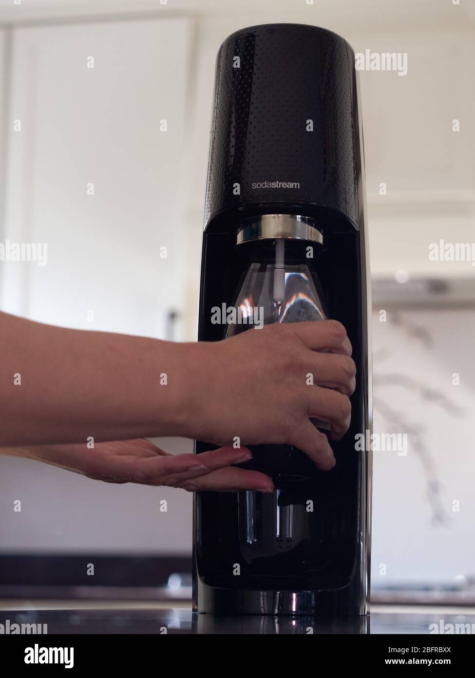 Non-caucasian woman using the Sodastream to carbonate water in a reusable plastic bottle in a modern kitchen with off white cabinets in the background Stock Photo