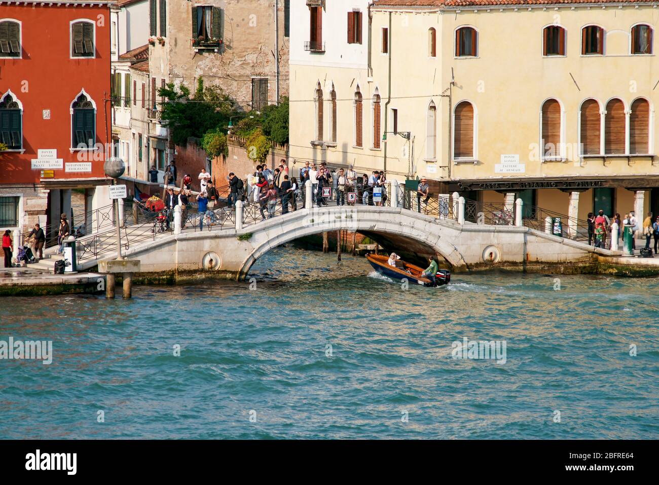 People standing on a bridge with a motorboat taxi passing under. Grand Canal into a small side canal between typical venetian style buildings.. Venice Stock Photo