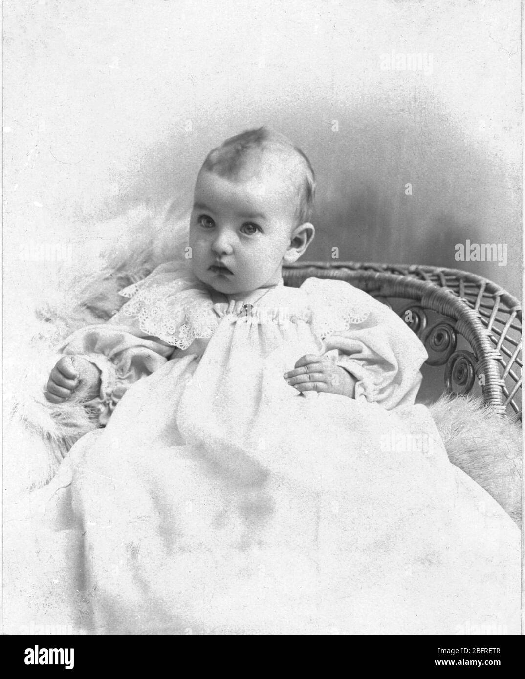 Wide-eyed, innocent baby in white, seated on a wicker chair.  c.1895.  To see my other vintage images, Search:  Prestor  vintage  kids Stock Photo