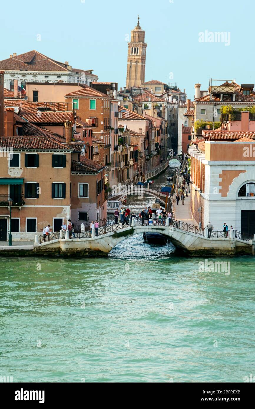 People standing on a bridge at the entrance to a small canal watching a ship pass by on the Grand Canal. The leaning bell tower of the church of San G Stock Photo