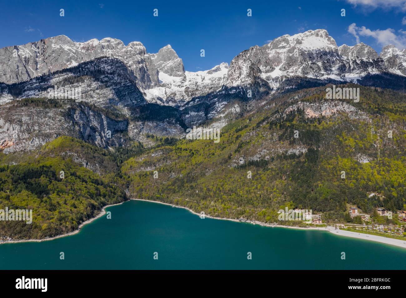 The Improbable aerial landscape of village Molveno, Italy, azure water of lake, empty beach, snow covered mountains Dolomites on background, roof top Stock Photo