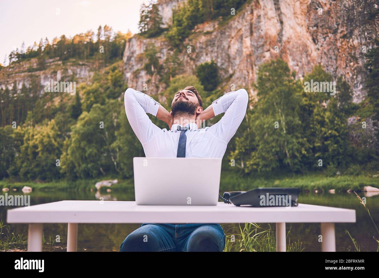 A bearded man works with a laptop on the nature. A happy freelancer is sitting and using an application or website on the grass. Work remotely in self Stock Photo