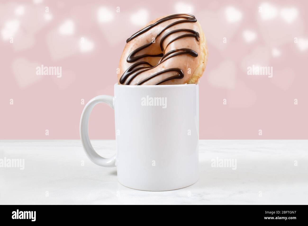 11 ounce white coffee mug filled with a glazed chocolate ice donut. Bokeh hearts glow in the pink background. Stock Photo