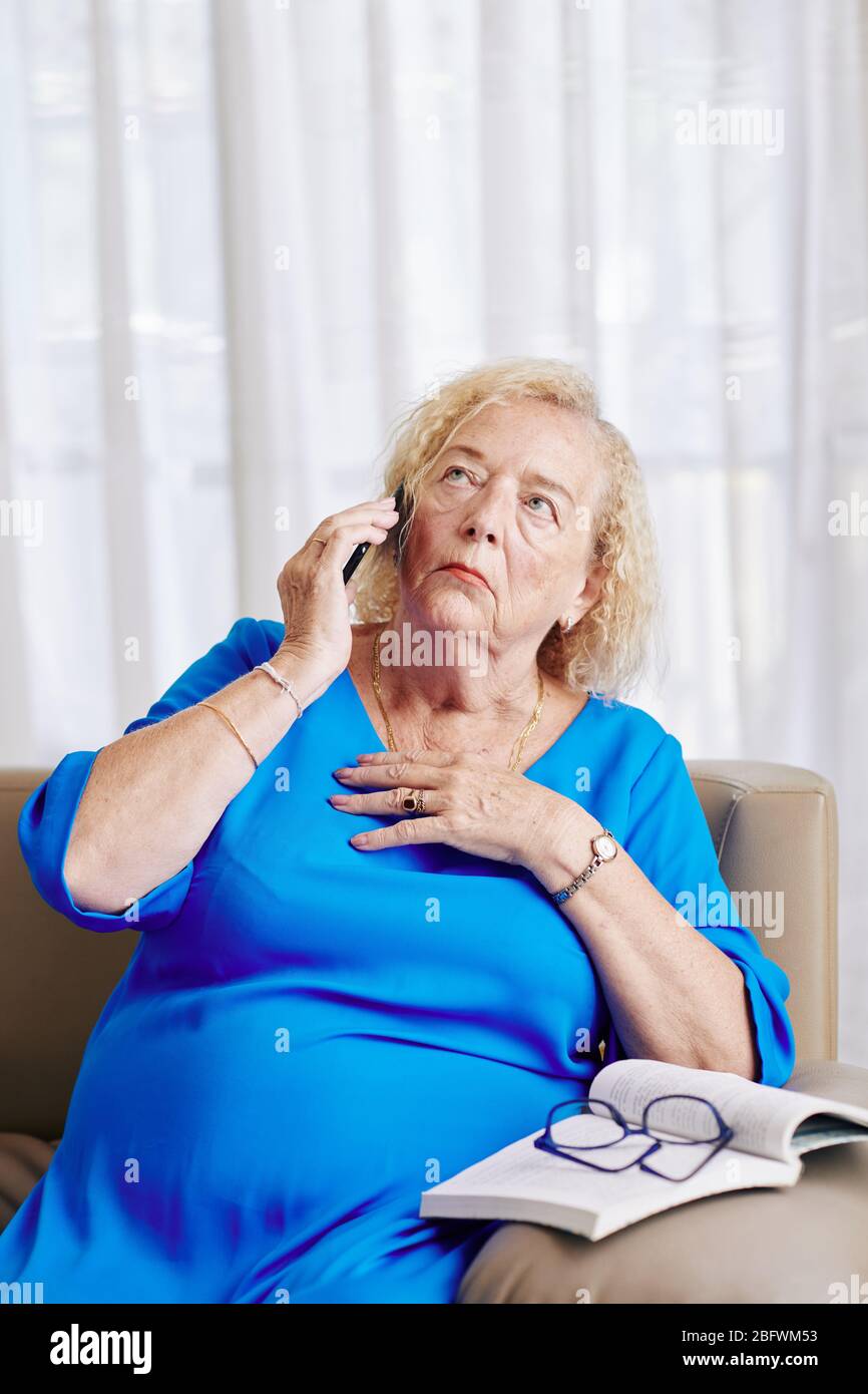 Nervous aged woman touching chest and looking up when talking on phone with family member Stock Photo