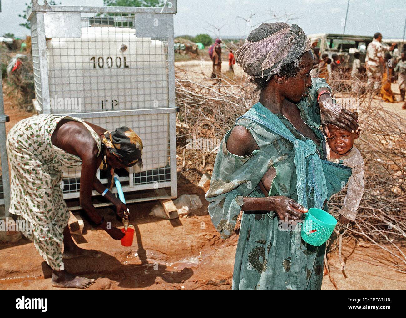 A Somali refugee woman holds her child as another woman gets water from a dispenser in the background at an aid station set up during Operation Restore Hope relief efforts. Stock Photo