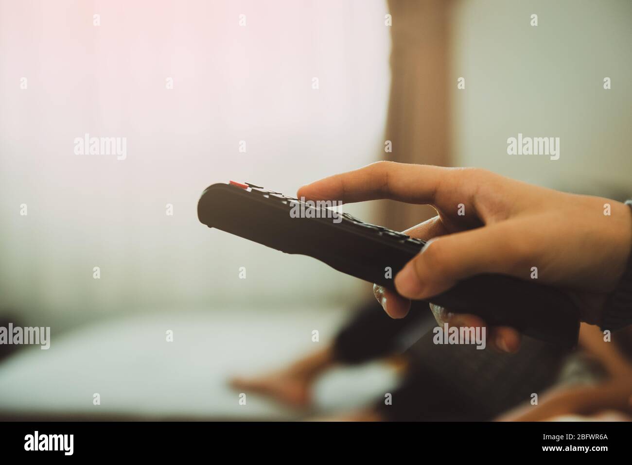 Close-up of young woman holding remote control and turning on the TV while sitting on sofa at home. Stop the virus Covid-19.Stay at home Concept. Stock Photo
