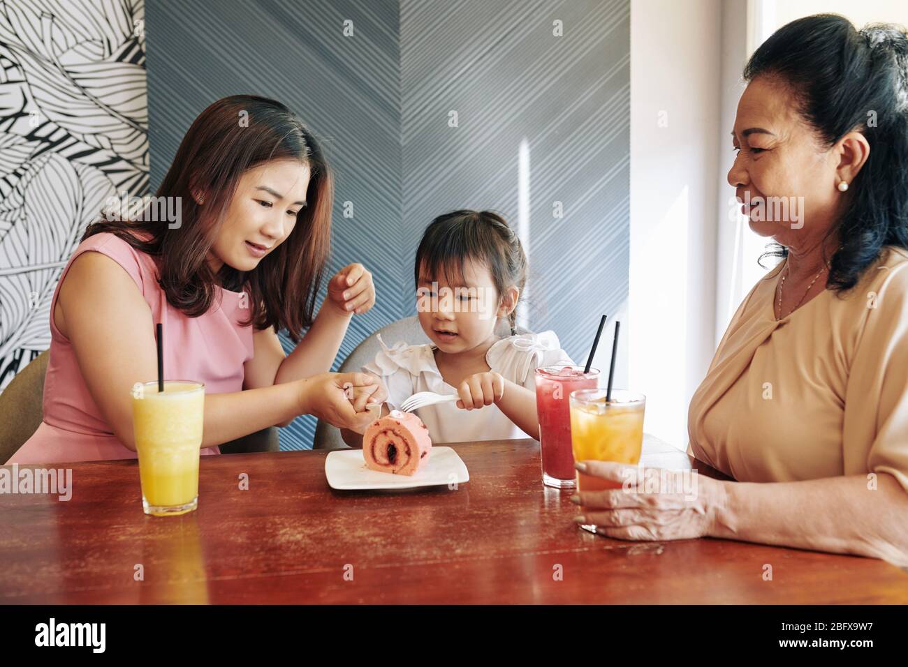 Young Vietnamese woman helping little daughter to cut a piece of sweet berry cake in cafe Stock Photo