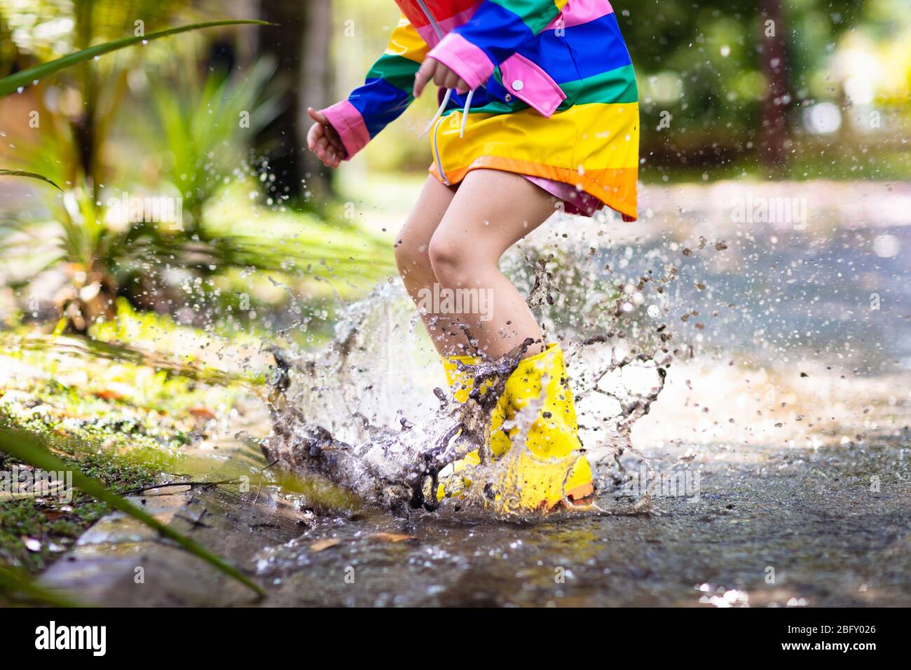 Kid playing in the rain in autumn park. Child jumping in muddy puddle on rainy fall day. Little girl in rain boots and rainbow jacket outdoors in heav Stock Photo