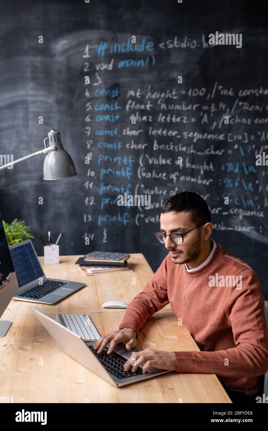 Talented young middle-eastern coder with beard sitting at desk and using laptop while working in office with blackboard Stock Photo
