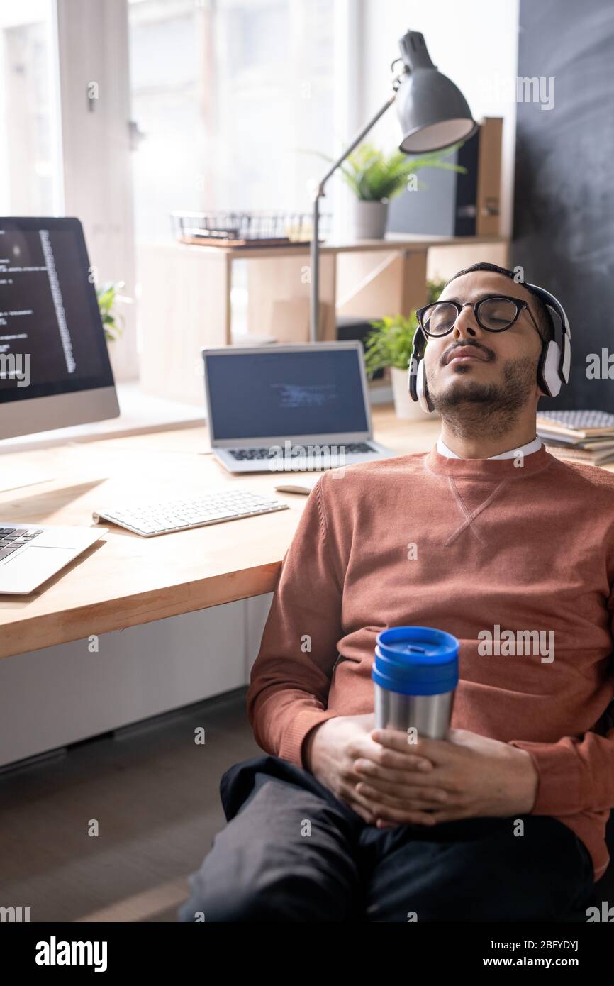 Exhausted young Arabian coder in headphones and glasses sleeping with thermos cup in hands at workplace Stock Photo
