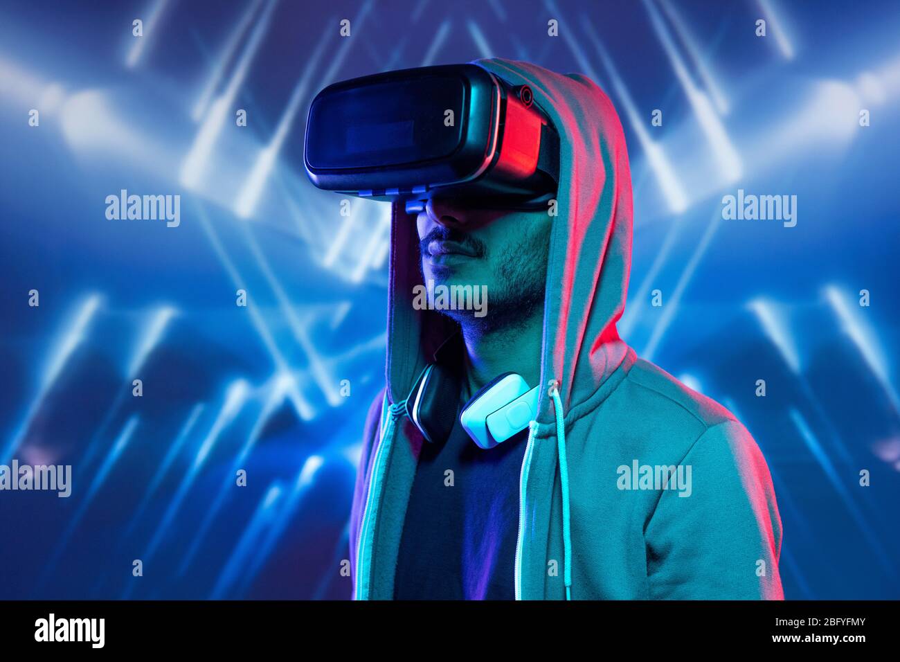 Hipster young man with stubble immersed in virtual reality wearing hoody sweatshirt watching 3D movie against blue luminous background Stock Photo