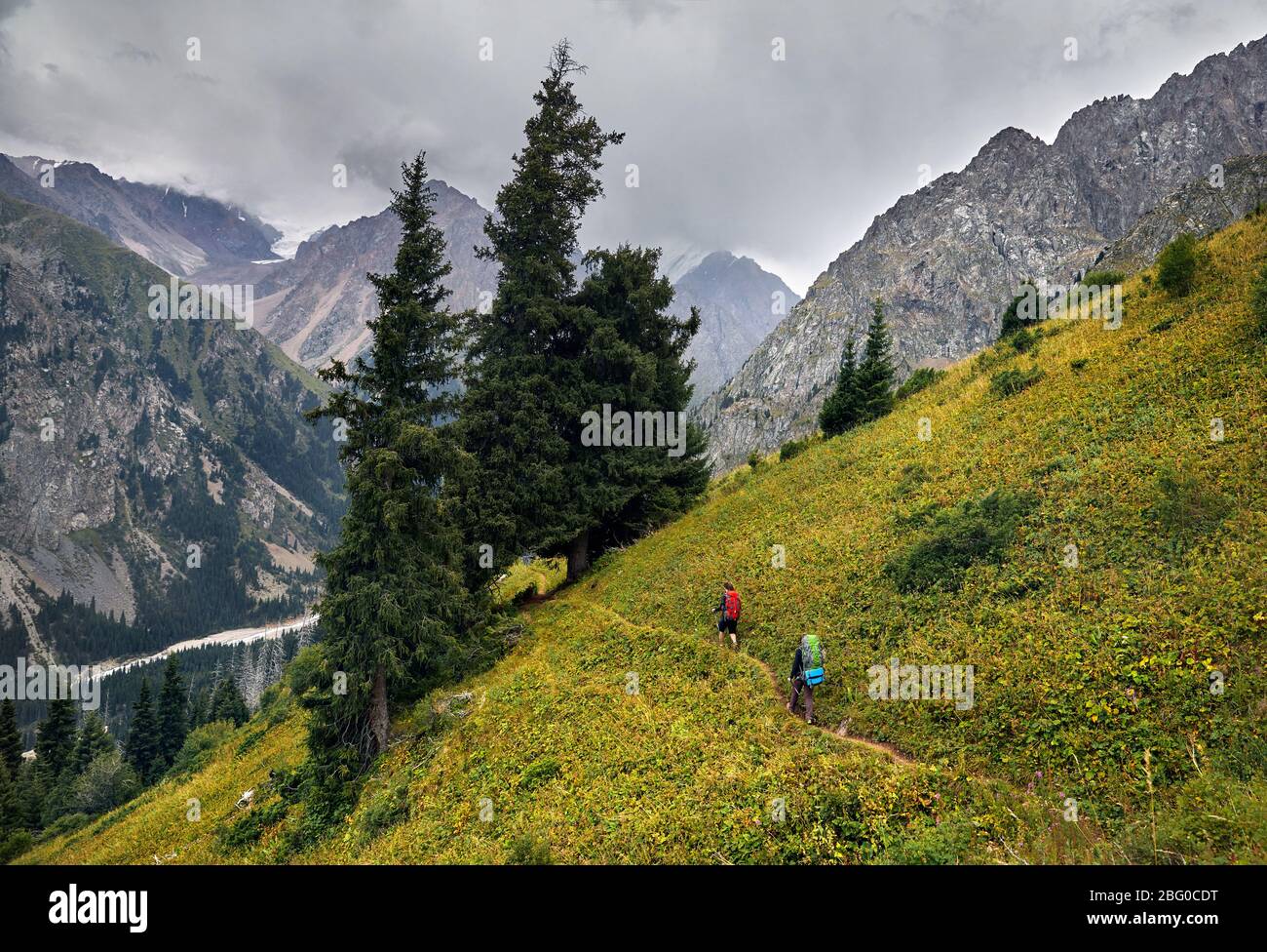 Two tourists with backpacks walking on the trail in the mountain valley at overcast sky background. Travel adventure concept Stock Photo