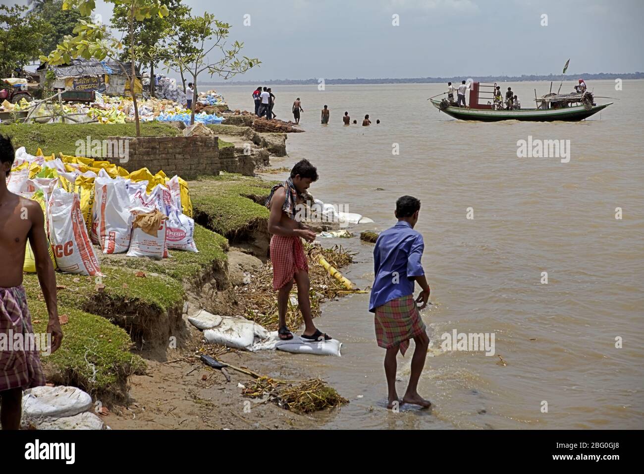Workers cleaning themselves on the bank of Rupnarayan river before lunch time during a river erosion control project, as a river crossing boat carrying passengers is arriving in the background in Tamluk, Purba Medinipur, West Bengal, India. Stock Photo