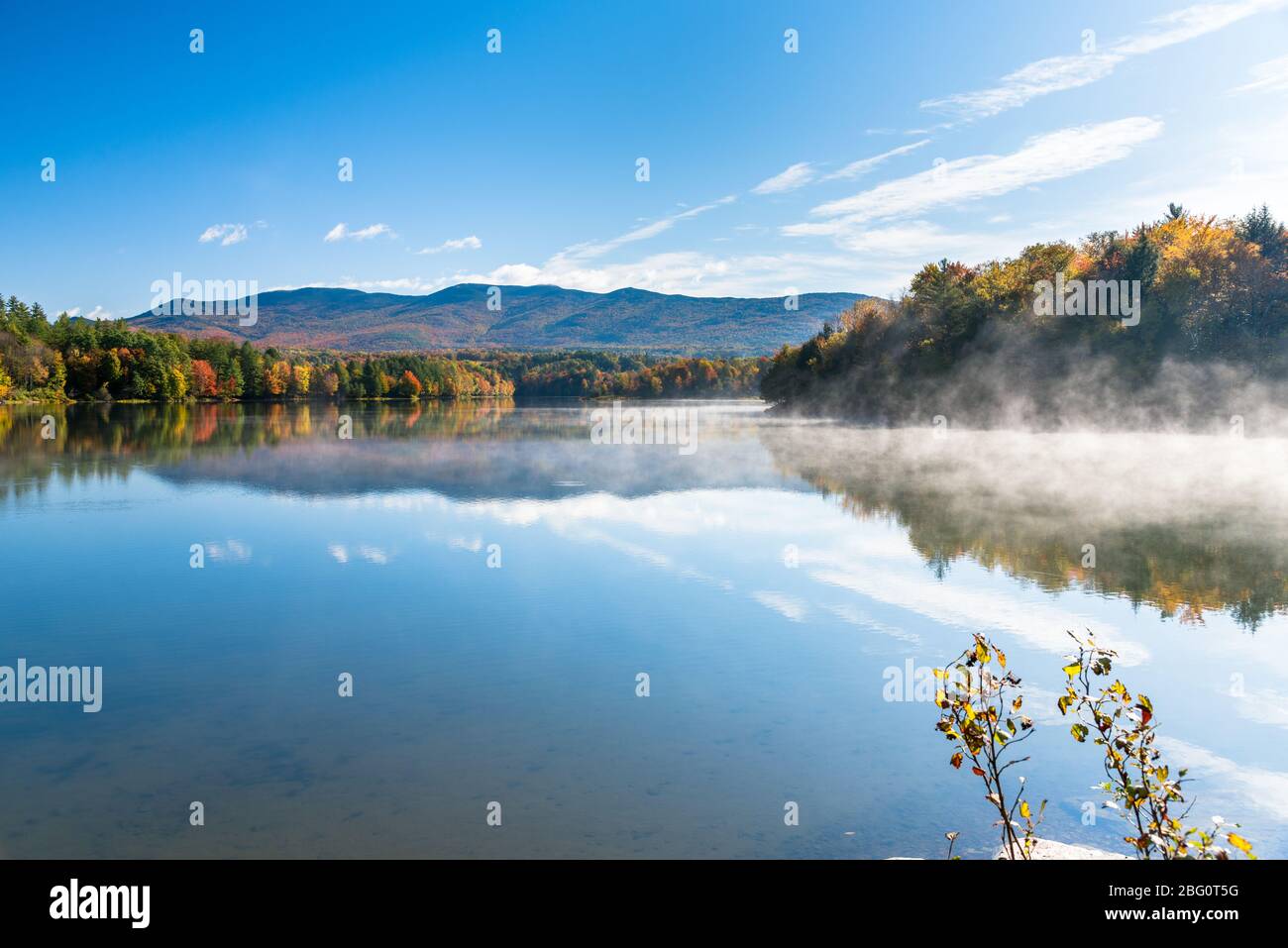 Lake with fog lifting from the water surface  in a magnificent forested mountain landscape on a sunny autumn morning. Stunniong fall foliage. Stock Photo
