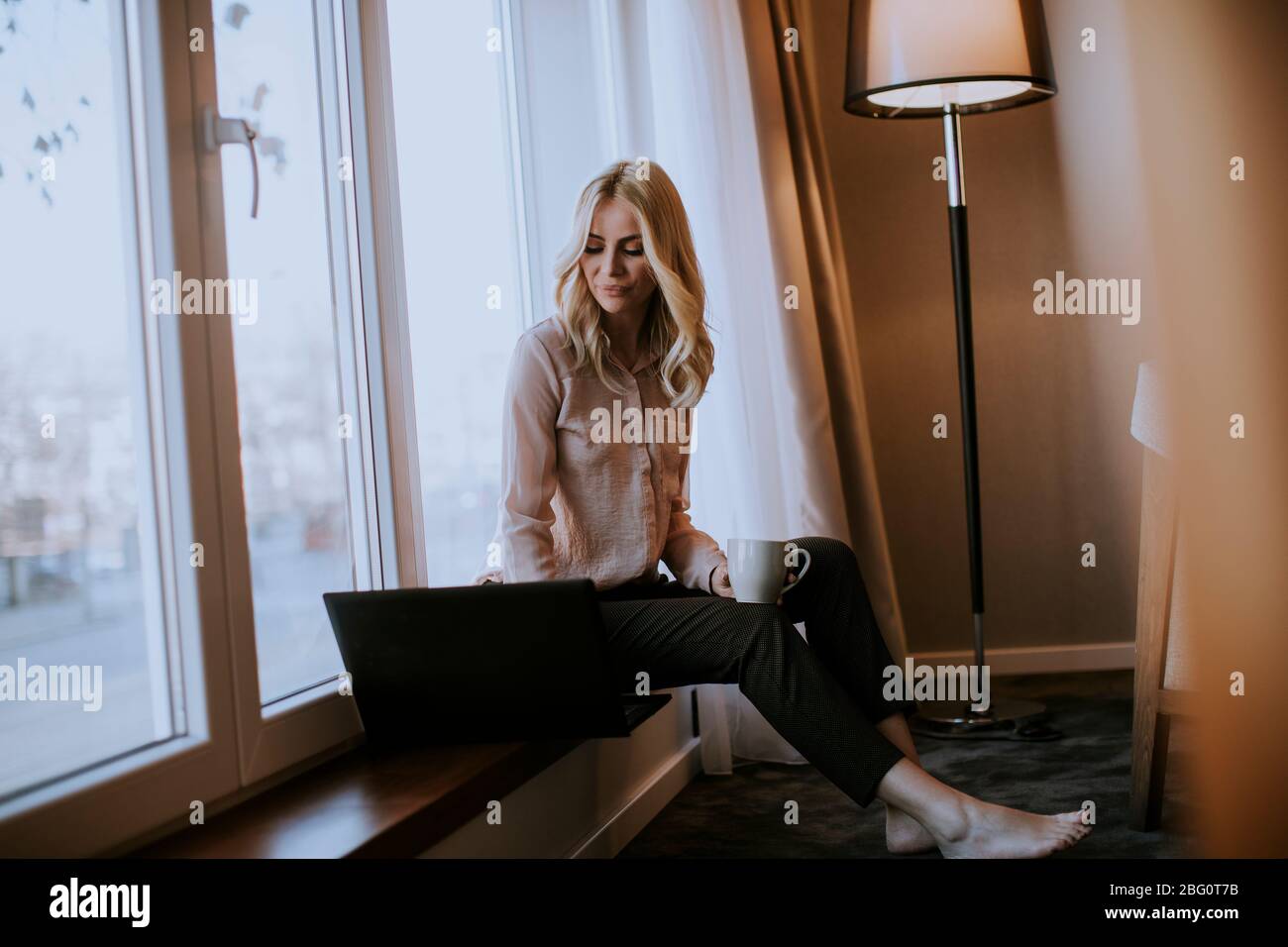 Pretty young woman working on laptop in living room on the window sill ...