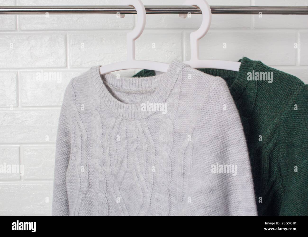 Clothes hang on hangers. Sweaters green and melange colors. Fashion and style, showcase, store, concept. White brick wall, comfort and cleaning, wool. Stock Photo