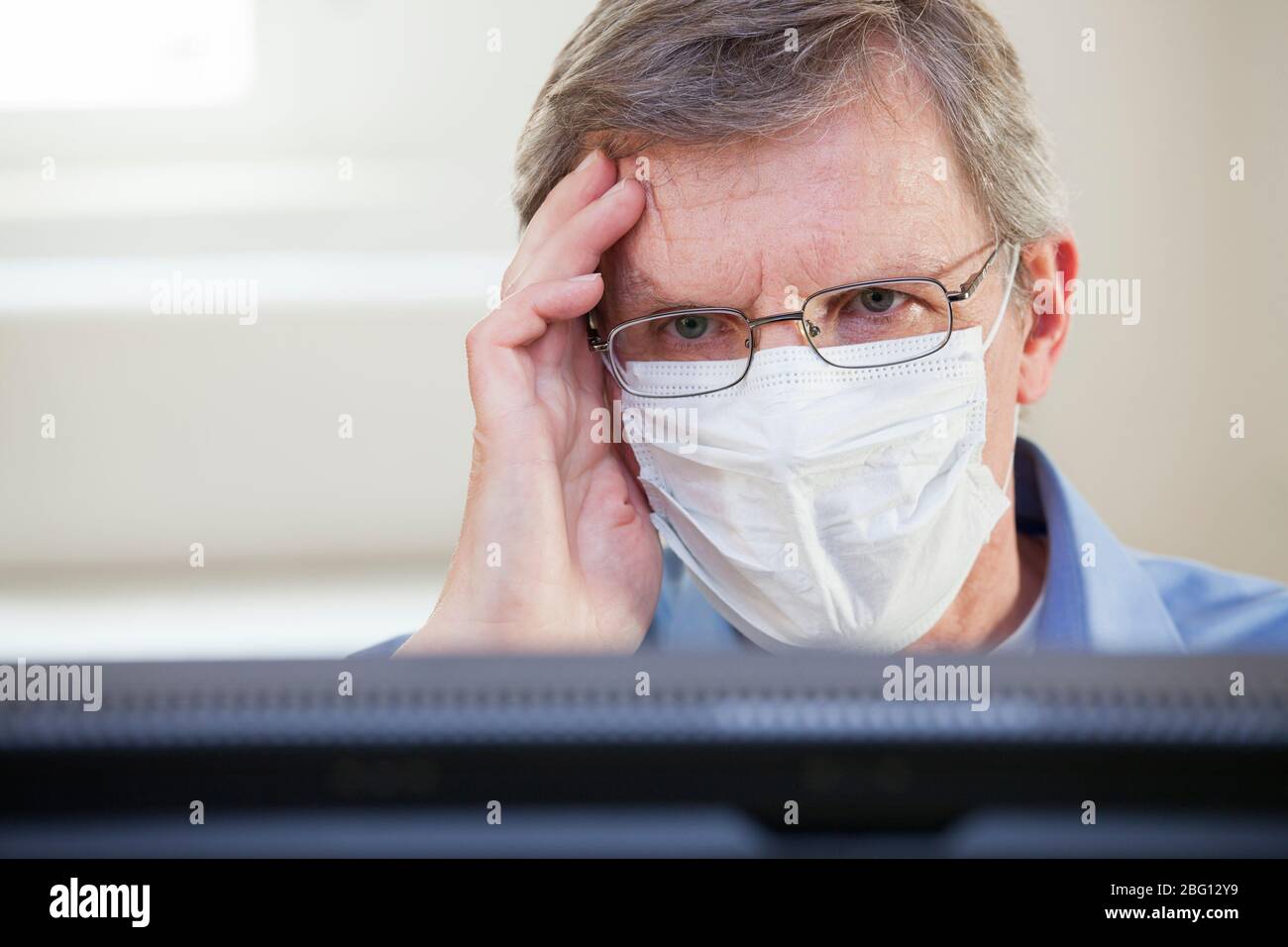 Sad businessman or doctor with face mask sitting behind a computer monitor in an office or  a hospital - selective focus Stock Photo